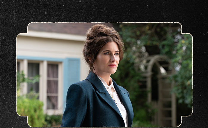 In a scene from Agatha All Along, Agatha Harkness (Kathryn Hahn) wears a navy blue fitted suit over a collared white shirt, and a gold pendant necklace. Her hair is in an updo, and the background features the exterior of a house and its garden.