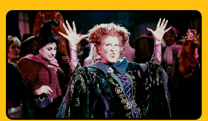 Winifred Sanderson from Hocus Pocus raising hands in spellcasting pose with crowd behind.