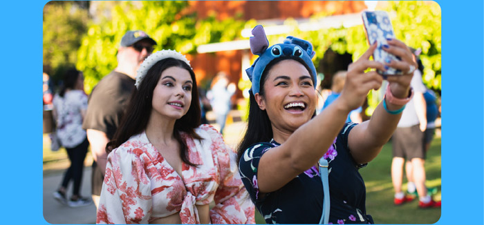 Two guests at a D23 outdoor event smile for a selfie-one in a floral top with a pearl-like headband, the other wearing a playful stick ear hat and holding up a phone to capture the moment.