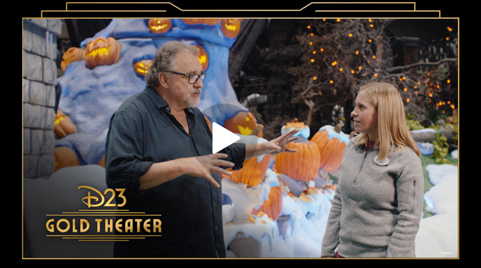D23 Gold Theater video still showing two people talking in front of of a Halloween-themed Disney set with glowing pumpkins.