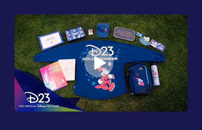 D23 Gold Member Collector set arranged on grass with a white play button overlay, showcasing Sorcerer Mickey spirit jersey, pin set, hat, backpack, water bottle, plaque, membership certificate, and collectible booklet.