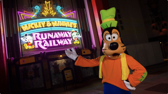 Goofy posing for a picture direction is arm towards a background of Mickey & Minnie’s Runaway Railway