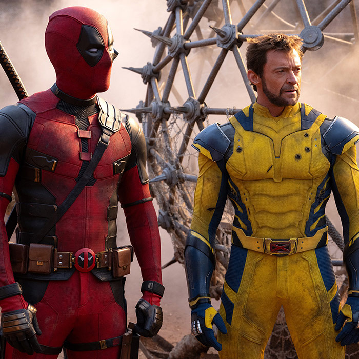 In a scene from Marvel Studios’ Deadpool & Wolverine, a masked Deadpool (left) wears a red and black suit next to Wolverine (right) who wears a blue and yellow suit that is scuffed and has several bullet holes in it. They are standing in a postapocalyptic landscape.