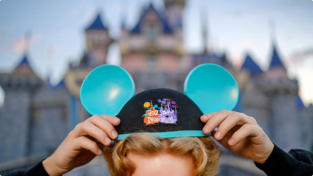 The image shows a close-up of a person adjusting their Mickey Mouse ear hat. The background is blurred, but silhouttte makes it clear that the location is in front of Sleeping Beauty Castle at Disneyland. The hat is black with cyan ears and trim, featuring an embroidered design with ‘D23 Day at Disneyland Resort’ in colorful text alongside images of iconic Disneyland symbols.
