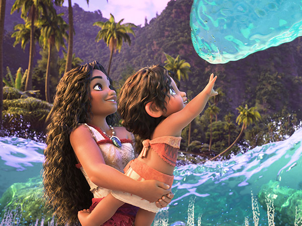 A scene from Moana 2: Moana is holding her little sister Simea up to touch a wave; they are standing on a Motunui beach, surrounded by the water.  In the background there are lush green mountains and palm trees.
