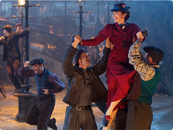 Mary Poppins, played by Emily Blunt, being lifted into the air by three singing lamplighters, she wears a red jacket and a navy blue hat with matching shoes, in a still from Mary Poppins Returns.