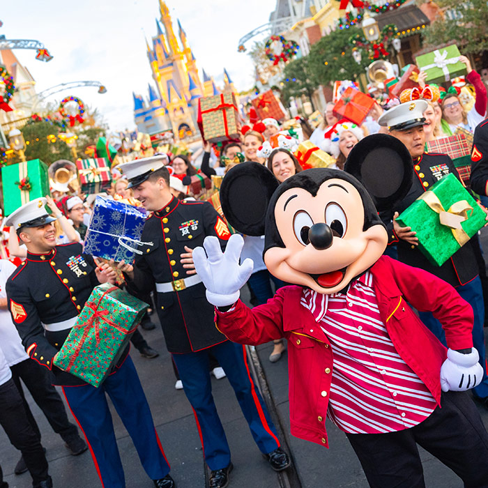 Mickey Mouse waves to the camera at Disneyland's Main Street, U.S.A., during the Mickey Around the World Toy Drive Tour. Behind him are Disney VoluntEARS and U.S. Marines, holding wrapped gifts and smiling.