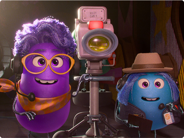 From Dream Productions, Paula (voiced by Paula Pell), a purple, anthropomorphic, jellybean-like director, wears a purple wig, orange glasses, and an orange necktie as she smiles at the camera from her director's chair. To her left is Xeni (voiced by Richard Ayoade), a turquoise, anthropomorphic, jellybean-like director with blue hair, brown glasses, and an orange striped turtleneck sweater. He also sits on a director's chair. The background features a buffet of food inside a bustling soundstage.