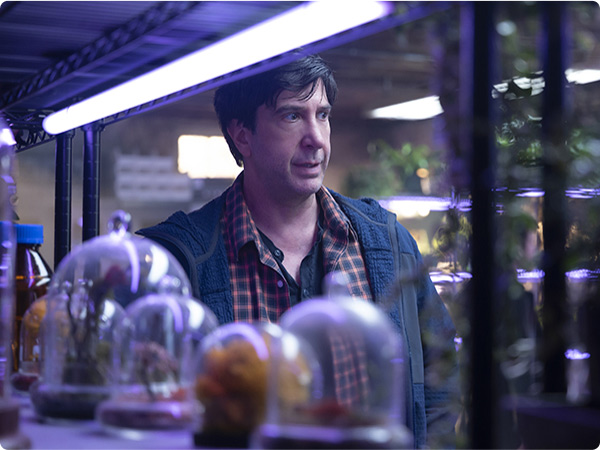 From Goosebumps: The Vanishing, Anthony Brewer (David Schwimmer) stands behind shelves of bell jars containing out of focus items. He is lit with purple lighting and looking towards the right of the frame.