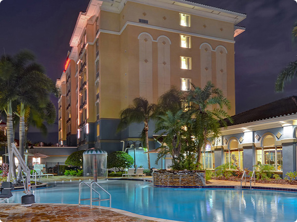 The Hilton Garden Inn Lake Buena Vista/Orlando sits brightly lit against the night sky. In front of the yellow hotel is the pool area. The pool is a turquoise blue color with the hotel lights reflected within the water. Multiple railings and steps are connected to the pool for access. Surrounding the pool are assorted lounge chairs and palm trees. Next to the pool is a small blue building with the lights on inside.