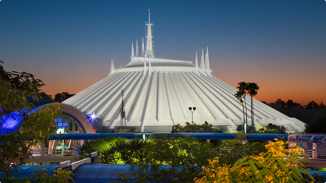 Disneyland's Space Mountain attraction illuminated during sunset, showcasing its iconic futuristic architecture with sharp spires and a sleek white dome. The structure stands against a vibrant orange and blue sky, surrounded by lush greenery.