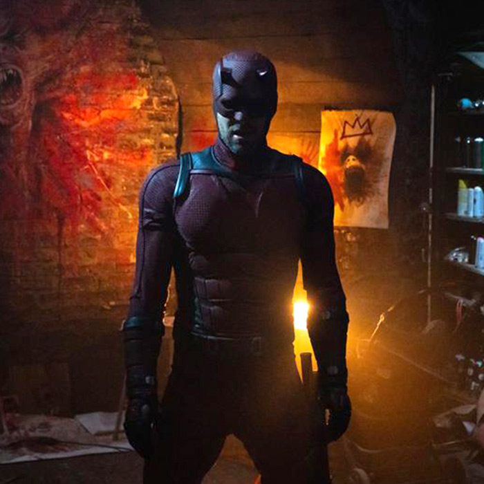 Daredevil stands in a dimly lit room with a fiery glow. The masked hero wears a dark armored suit, with a graffiti crown on a wall behind him.