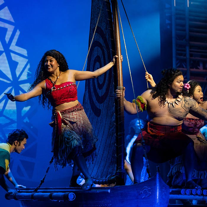 Live-action Moana stage performance, Moana stands confidently on a boat, surrounded by Polynesian performers in a dynamic stage setting.