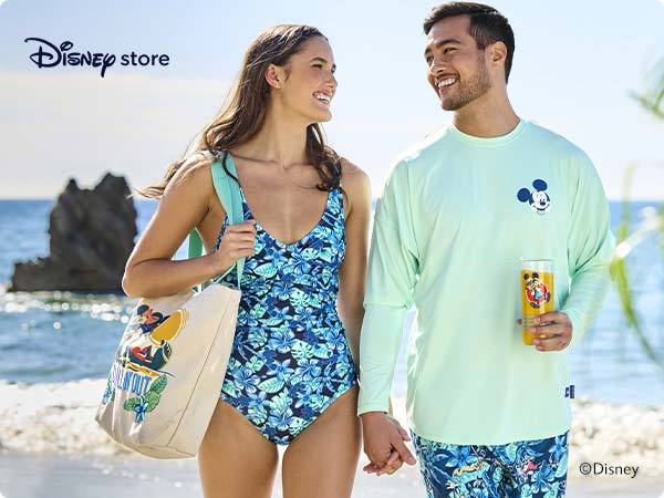 Disney Store beachwear collection. A smiling couple in matching Mickey-themed beachwear walk hand in hand by the ocean.