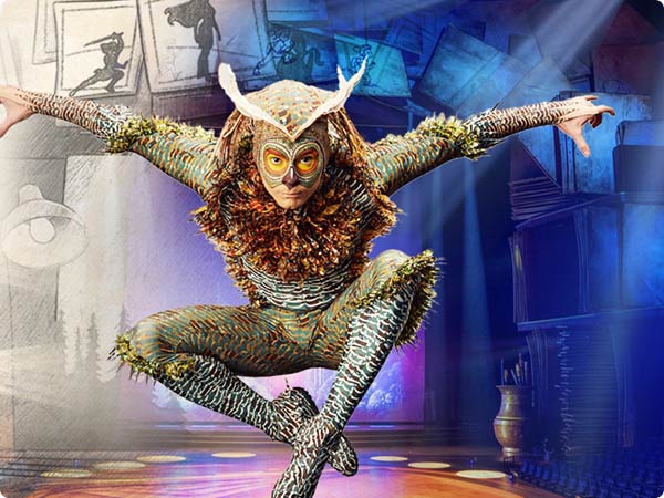 Performer in owl-themed costume leaps midair on a spotlighted stage in 'Drawn To Life' by Cirque du Soleil