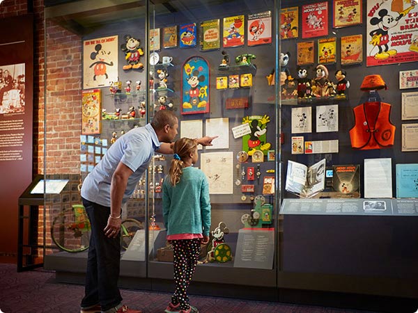 Adult and child point at a glass display filled with vintage Mickey Mouse toys, books, and collectibles inside a museum exhibit.