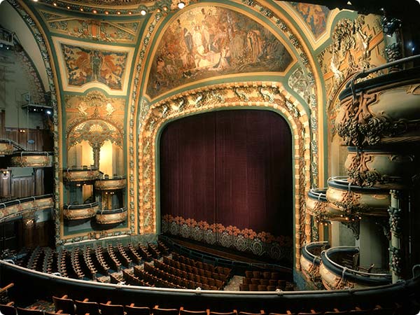 Lavishly decorated theater with ornate gold detailing, balconies, and a red velvet curtain center stage.