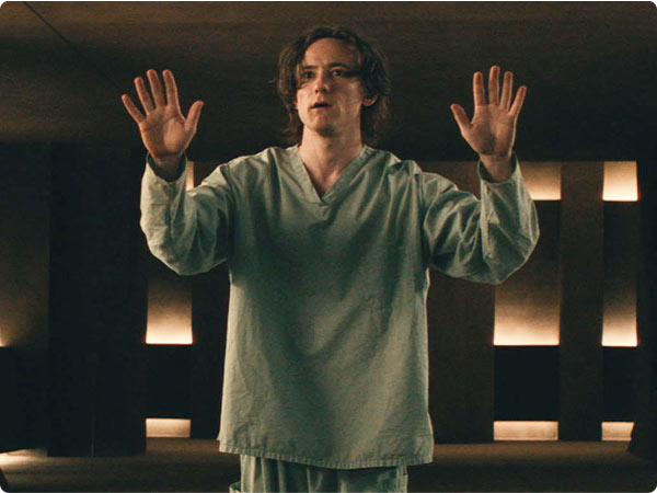 Disheveled man in mint scrubs raises hands inside dim modern chamber lit by narrow wall lights.
