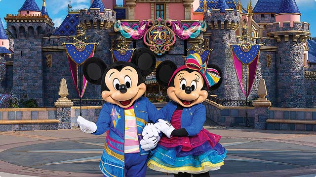 Mickey and Minnie in vibrant 70th-anniversary outfits pose before a richly decorated Sleeping Beauty Castle.
