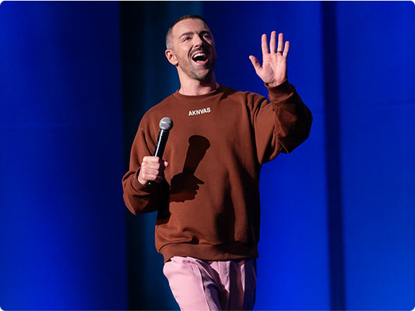 Comedian Matteo Lane in a brown sweater, waving while holding a mic on a blue-lit stage.