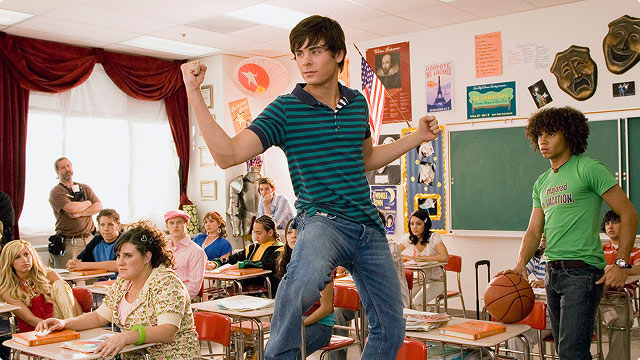 High School Musical classroom scene- Troy Bolton strikes a pose on a desk while classmates look on.