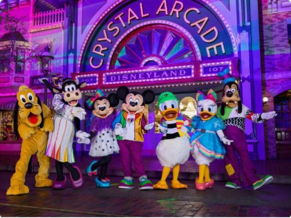 Pluto, Clarabelle, Minnie, Mickey, Donald, Daisy, and Goofy pose in bright outfits outside Disneyland's Crystal Arcade at night.