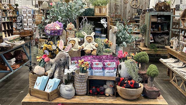 Rustic shop table packed with plush bunnies, teddy bears in hoodies, blooms, and purple snack bags labeled “POPPY Spring Confetti”