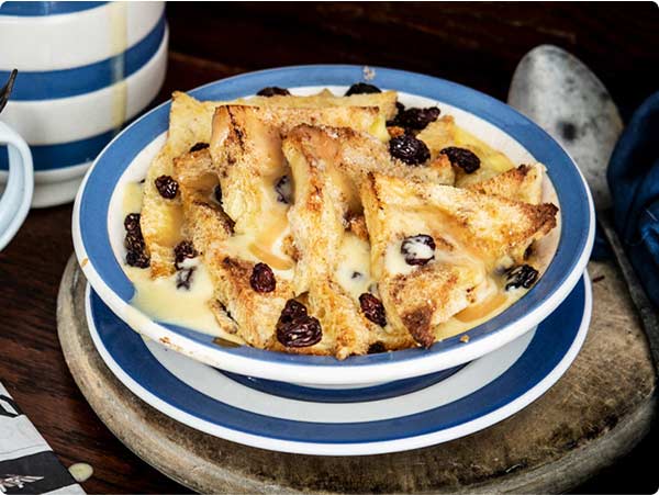 Raglan Road’s Famous Ger's bread and butter pudding sits on a table, housed in a white and blue bowl. The dish is served warm with a hot butterscotch sauce with cool creme anglaise in a cup off to the side.