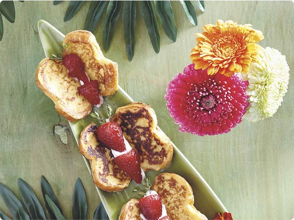 Three Mickey-shaped French-toast bites with strawberries on a green platter next to vivid pink, orange and white blooms.