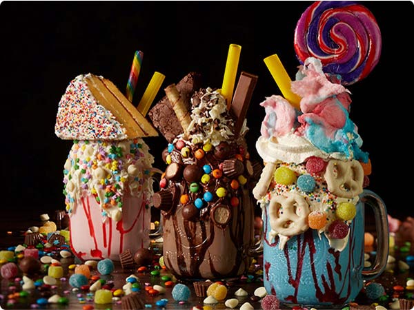 Several Super Nova Shakes from Planet Hollywood: one sprinkle-rimmed strawberry shape topped with a waffle slice and rainbow candy, a chocolate shake pilled with brownies, popcorn and mini peanut-butter cups, and a blue cotton-candy shake crowned with a swirl lollipop-each shake jar is overflowing onto a tabletop covered in colorful sweets.