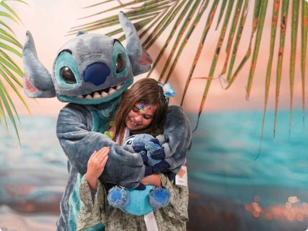 Tropical setting: costumed Stitch hugs a smiling girl, with palm fronds framing a turquoise-blue ocean backdrop.