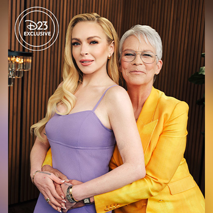 Jamie Lee Curtis embraces Lindsay Lohan in front of wood paneling; Lohan wears a lavender dress beneath a “D23 Exclusive” logo.