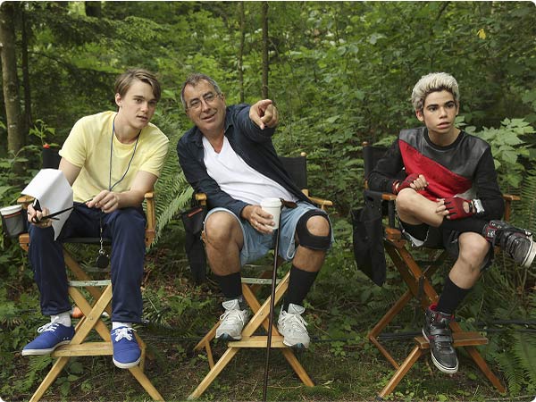 Mitchell Hope, Kenny Ortega, and Cameron Boyce sit on director chairs in a forest while filming Descendants (2015).
