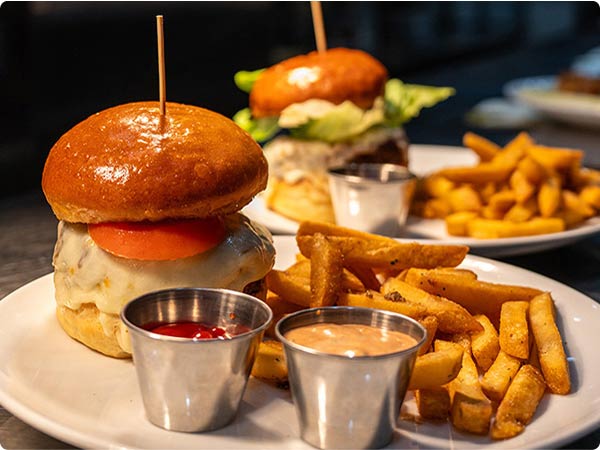 Two cheeseburgers with fries and dipping sauces on white plates, set under warn lighting on a dark table.
