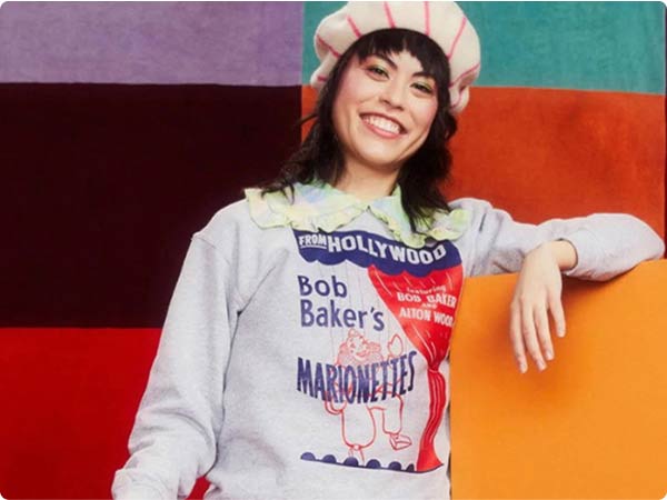 Woman in Bob Baker’s Marionettes sweatshirt and striped hat smiling against a colorful background.