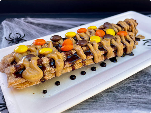 The'Spooky Churro' a seasonal treat drizzled with chocolate and peanut butter topped with colorful candy pieces on Halloween-themed plate.