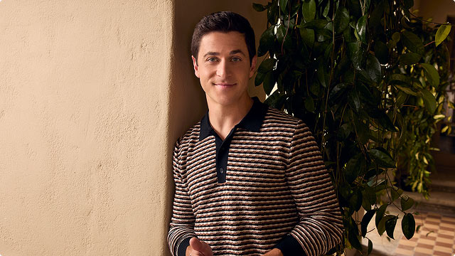 Justin Russo (played by David Henrie) smiling in striped shirt standing beside a wall with plants in the background.