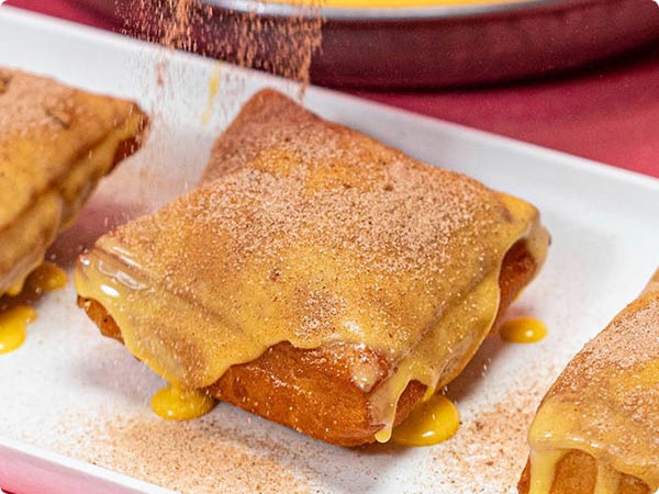 A warm golden-brown beignet, covered in a sweet yellow syrup, and is being topped with brown sugar. The treat rests on a white plate with two identical beignets on either side of it.
