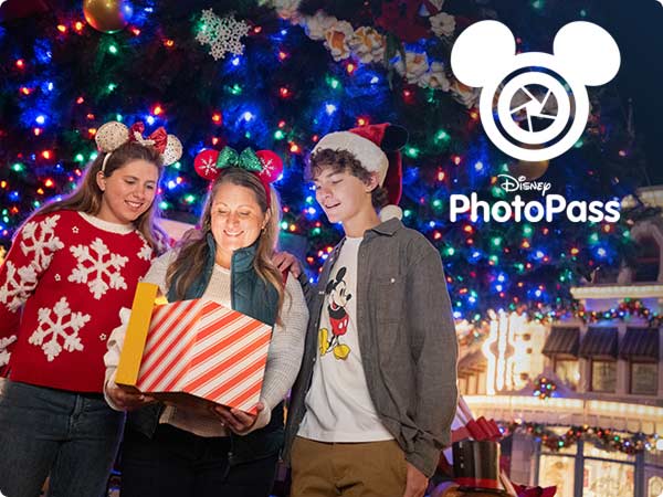 Group of guests in holiday outfits posing with a wrapped gift in futon of a brightly lit Christmas tree, promoting Disney PhotoPass.