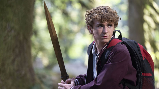 Percy Jackson stands in a forest holding his sword, looking alert with a backpack over one shoulder.