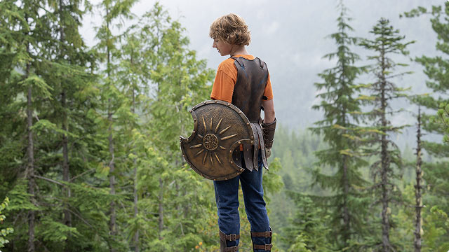 Percy Jackson standing in a forest clearing wearing armor and holding a sun-emblem shield.
