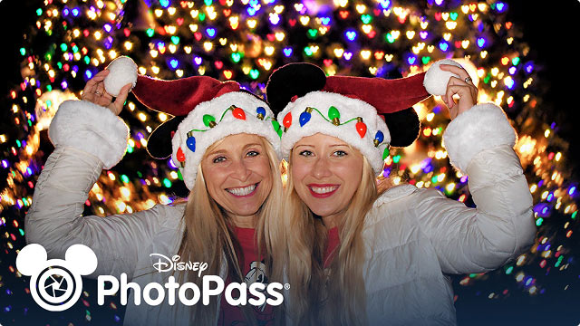 Two guests wearing matching Mickey holiday Santa hats posing in front of colorful holiday lights with the Disney PhotoPass logo.