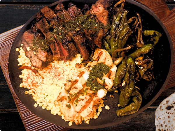 A full Ribeye steak with grill marks, two elotes corn cobs and a medley of green peppers on a light blue plate served on a dark wooden serving paddle. The golden Maya Grill logo sits in the top left.