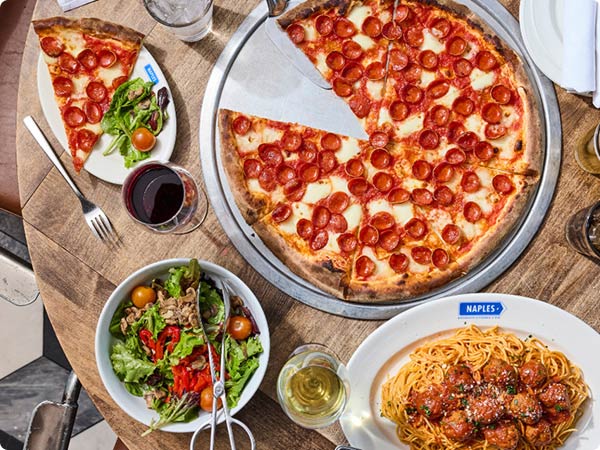 A circular wooden dining tabled filled with an Italian meal from Naples Ristorante including: A huge pepperoni pizza, spaghetti and meatballs, salad and small dish of parmesan cheese, and assorted beverages.